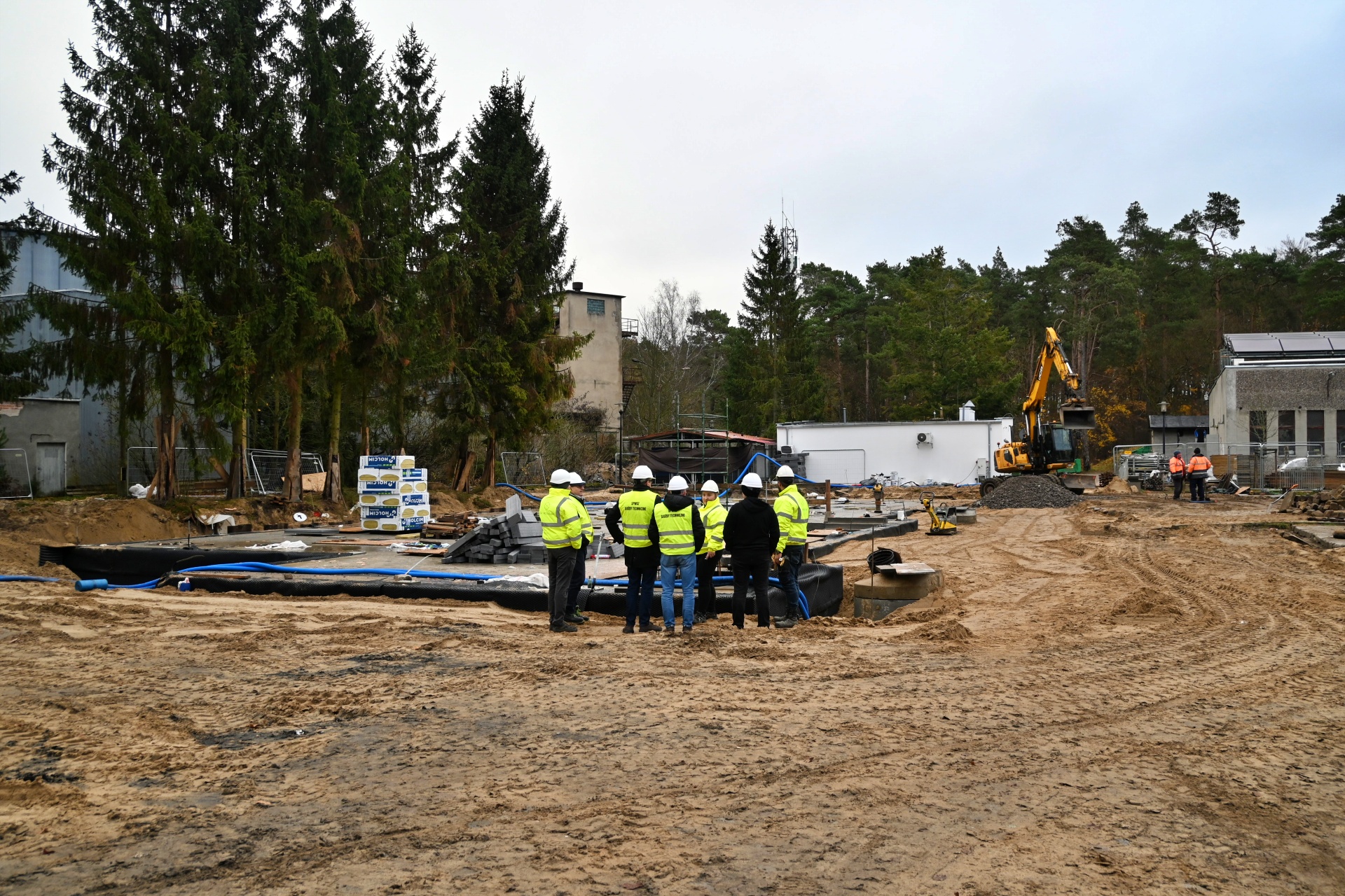 Plac budowy - grupa osób ubrana we fluorescencyjne kamizelki z kaskami budowlanymi na głowach stoi na piachu przy płycie fundamentowej.