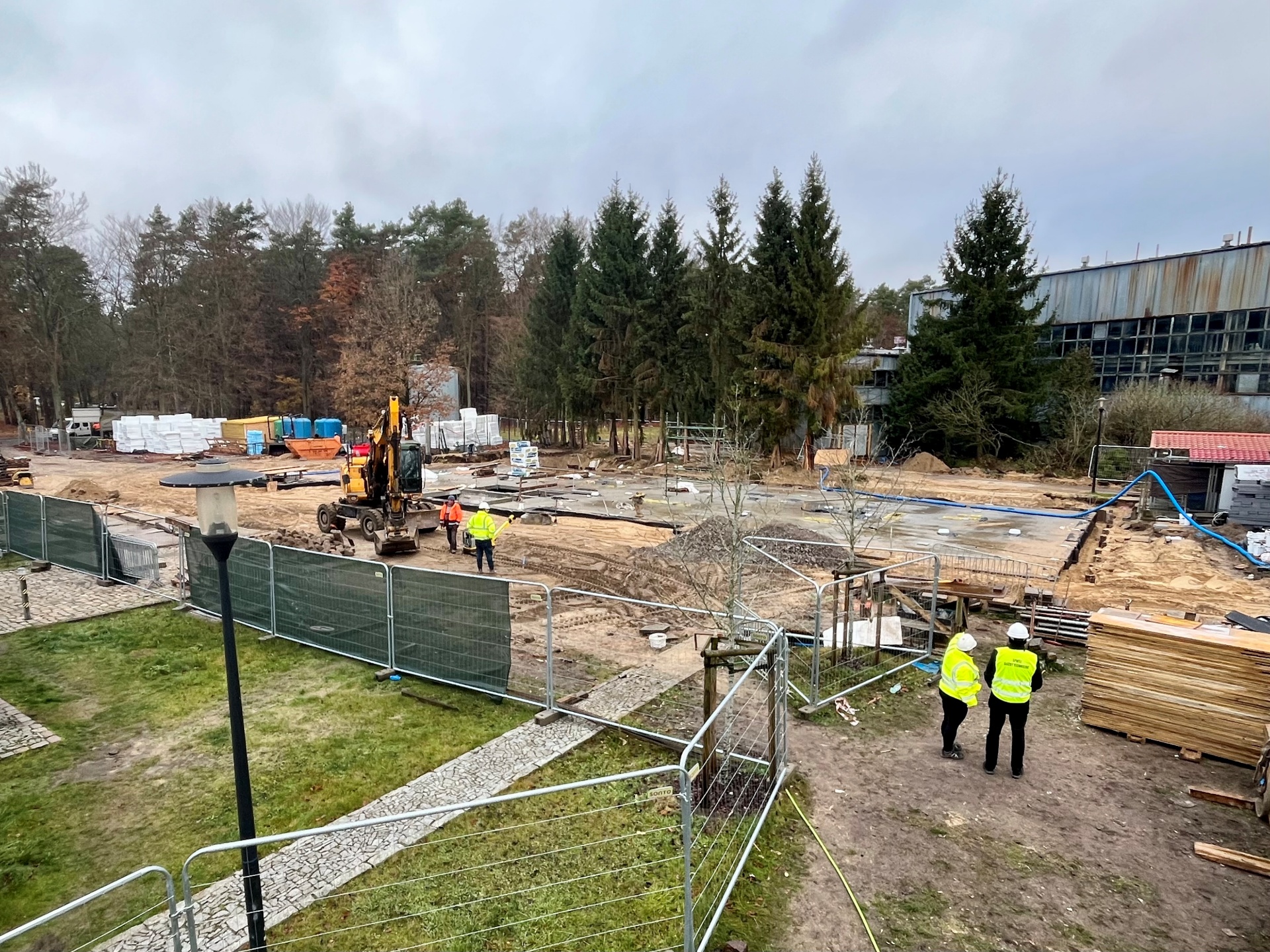 W centralnym punkcie fotografii widoczna płyta fundamentowa nowego budynku. Teren budowy ogrodzony płotem. W kilku miejscach osoby ubrane we fluorescencyjne kamizelki z kaskami budowalnymi na głowach.